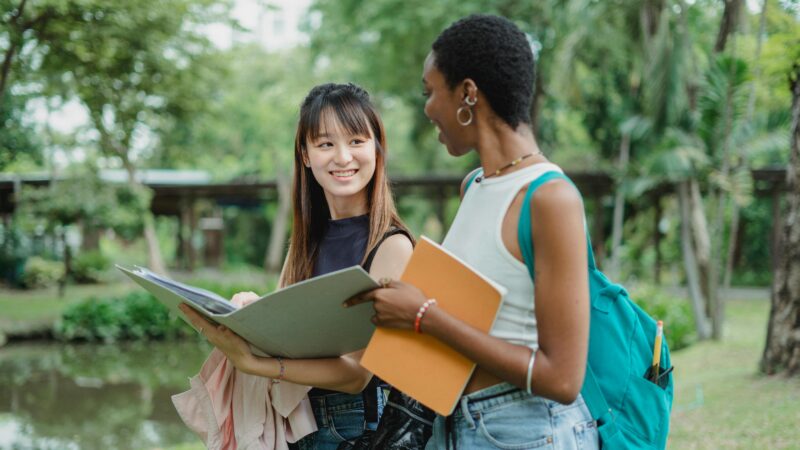 Multiethnic female friends speaking while walking in park with notepads